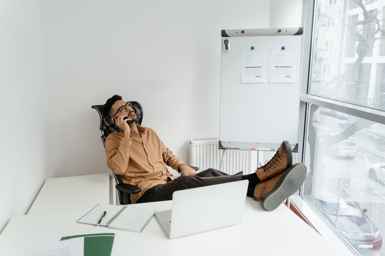 A man in a brown shirt sitting and talking on the phone, caught in the 'I'll do it later' trap that delays important tasks and ruins productivity.