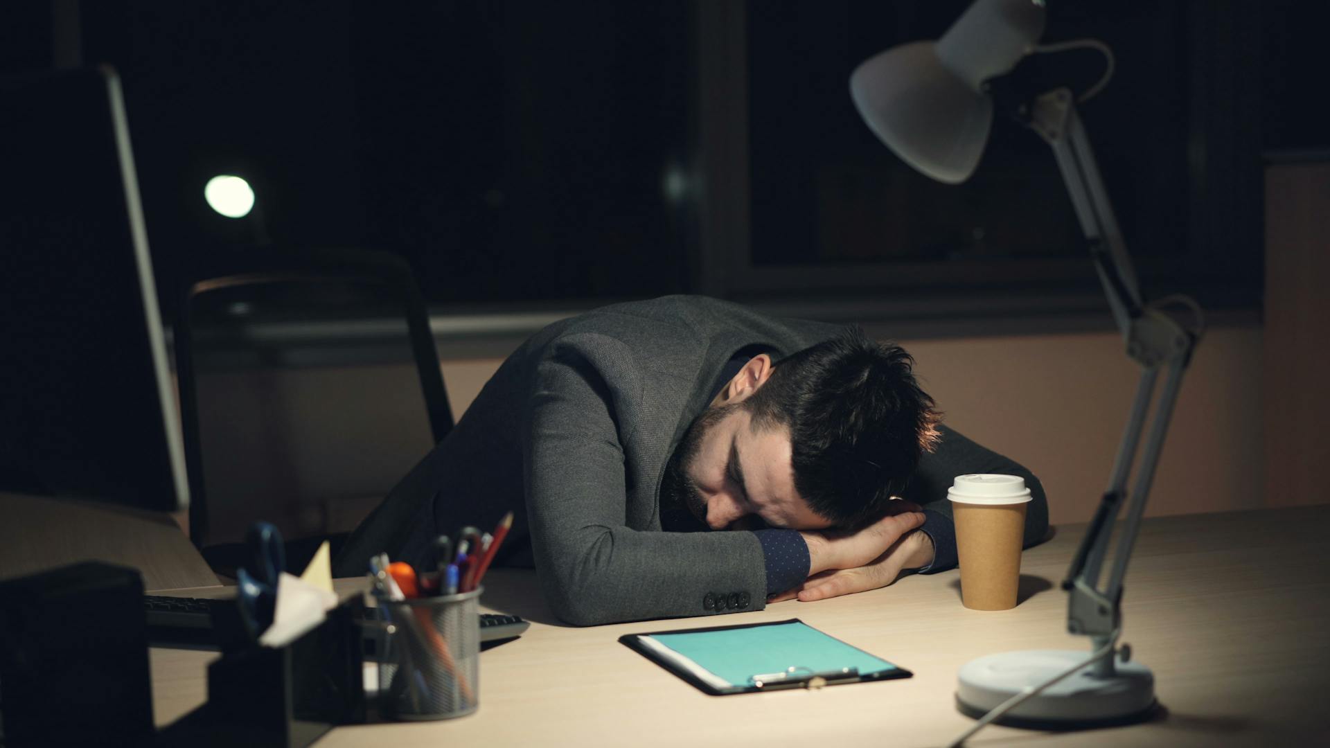 Exhausted office worker asleep at desk with laptop and coffee, showing burnout.